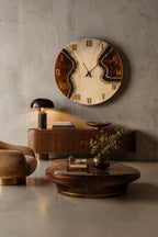A large circular wall clock featuring a cream-colored center with dark brown resin edges and black crystal accents. The clock face displays gold Roman numerals and thin black hands, mounted on a wall above a wooden sideboard and coffee table.