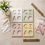 Four square tiles in white, sage green, dusty pink, and pale yellow, each featuring the cutout letters C, I, A, and O. The set includes a glass vase with white tulips, several folded silk fabric squares, and two gold-toned spirit levels.