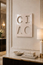 A square, glossy, off-white ceramic wall plaque featuring the debossed letters C, I, A, and O arranged in a two-by-two grid. The surface has a smooth, reflective finish with subtle indentations forming the characters.