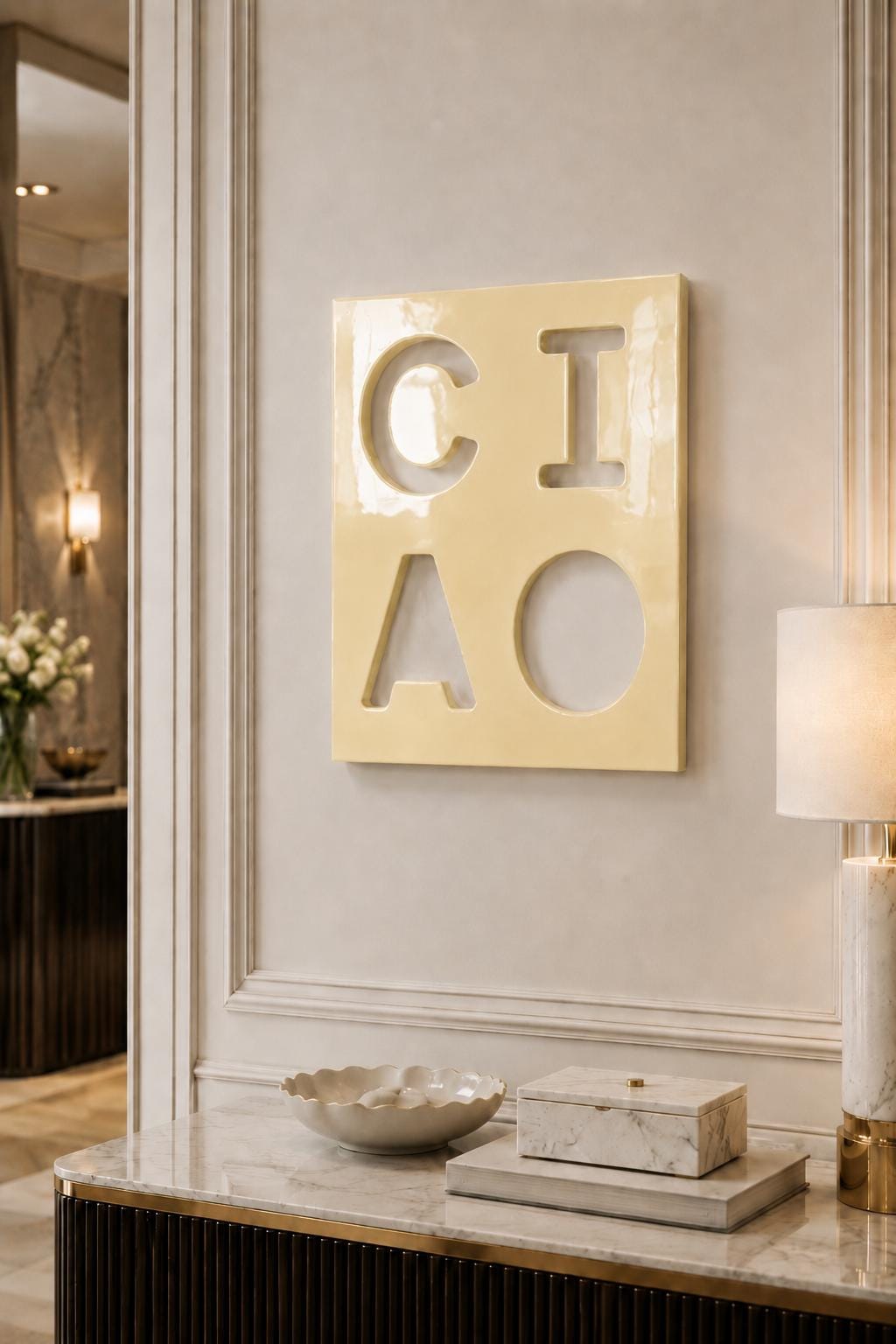 A square, glossy, pale yellow wall plaque featuring the word CIAO cut out in a bold, block-letter font. The letters C, I, A, and O are arranged in a two-by-two grid, revealing the wall surface behind the smooth, reflective surface.