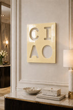 A square, glossy, pale yellow wall plaque featuring the word CIAO cut out in a bold, block-letter font. The letters C, I, A, and O are arranged in a two-by-two grid, revealing the wall surface behind the smooth, reflective surface.