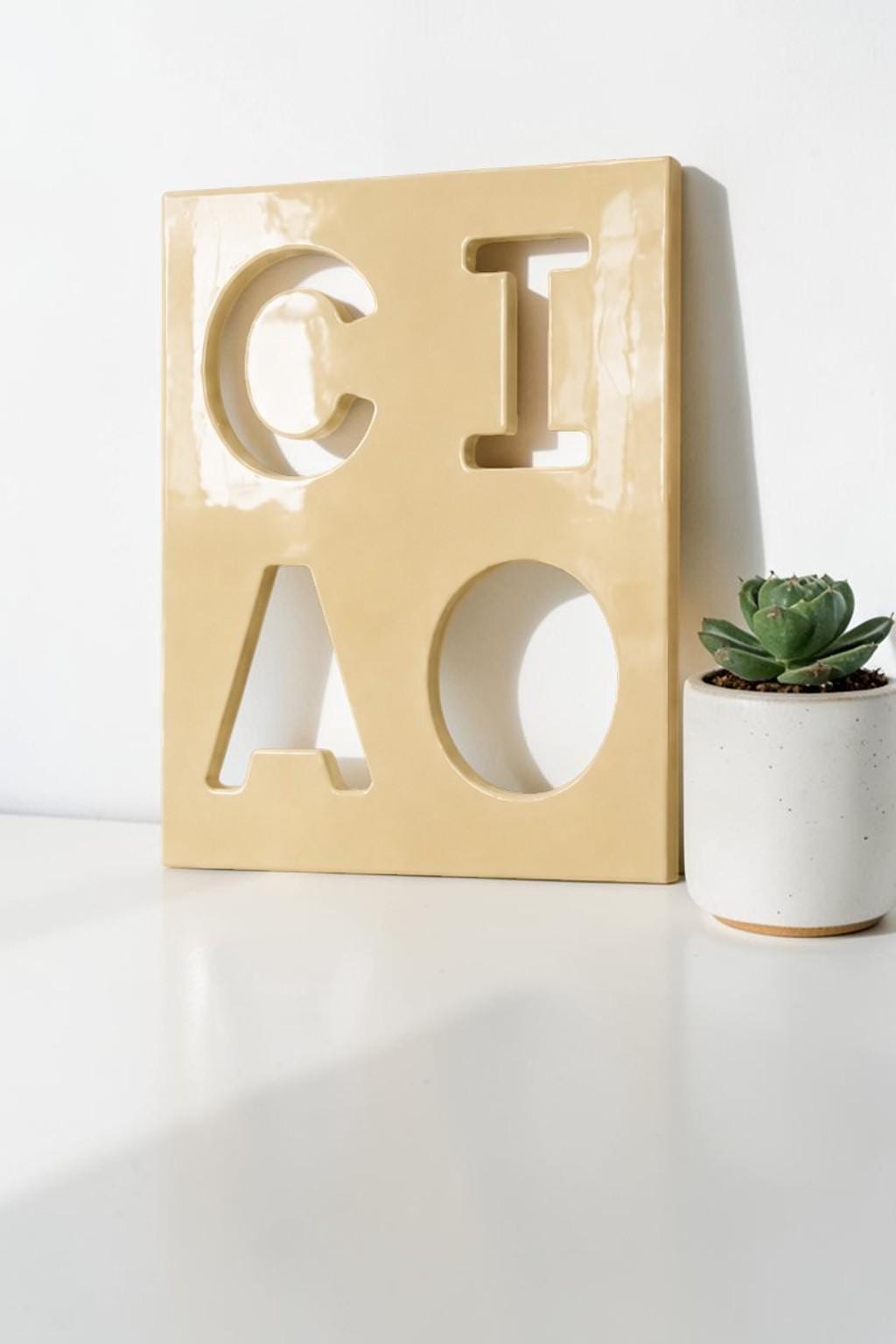 A square, glossy, light yellow ceramic plaque featuring the cutout letters C, I, A, and O arranged in a two-by-two grid. The letters are negative space cutouts within the solid, smooth, light yellow surface of the ceramic tile.