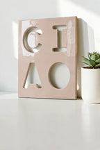A rectangular, glossy, light pink ceramic wall plaque featuring the cutout letters C, I, A, and O arranged in a two-by-two grid. The surface has a smooth, reflective finish with visible light reflections across the front face.