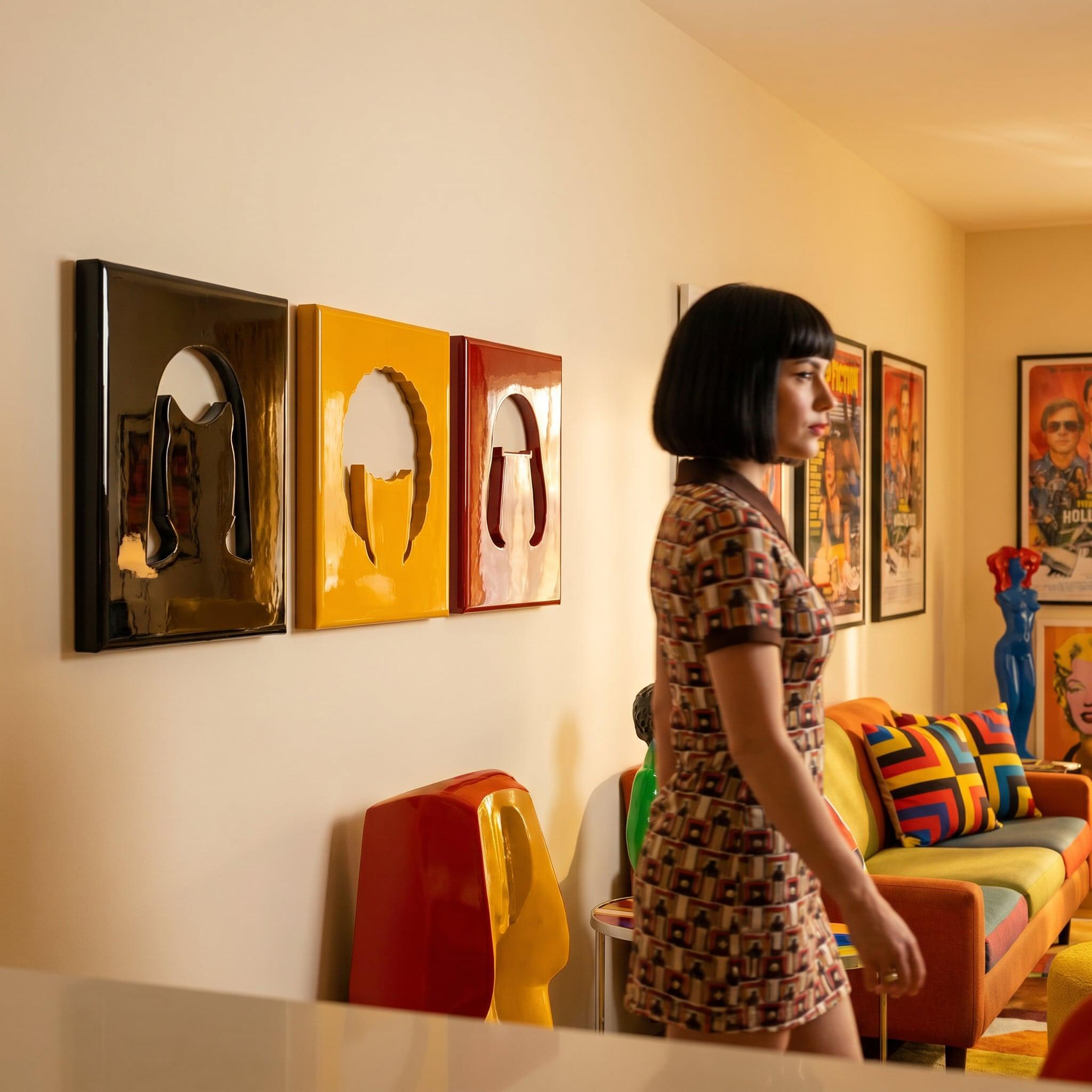Three rectangular wall art pieces in black, yellow, and red, each featuring a central cutout silhouette. A person with a dark bob haircut wears a short-sleeved patterned dress, standing near a colorful sofa with geometric cushions.