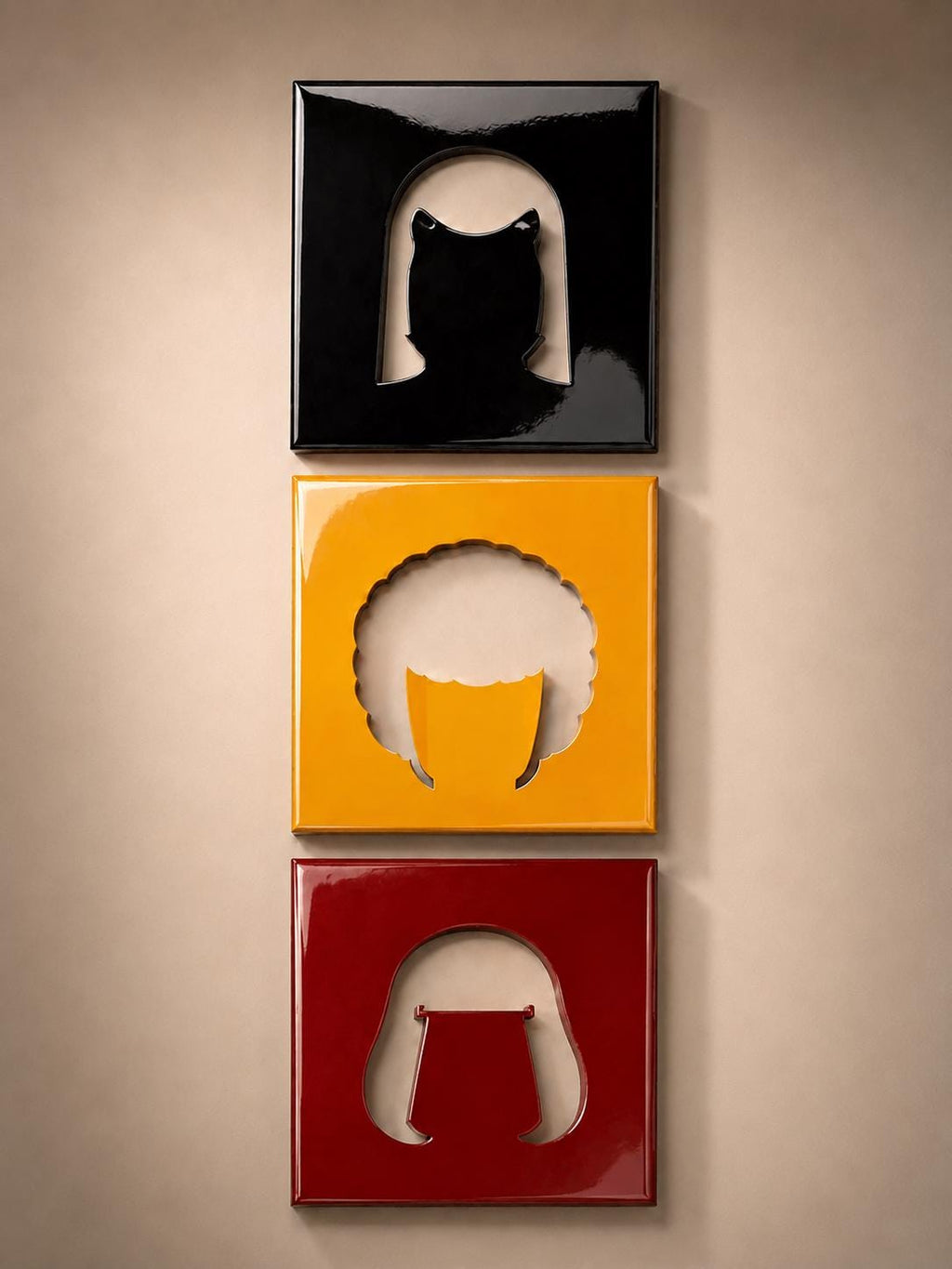 Three square wall plaques arranged vertically. The top plaque is glossy black with a cutout silhouette of a bob hairstyle. The middle plaque is glossy yellow with a cutout of an afro hairstyle. The bottom plaque is glossy dark red.