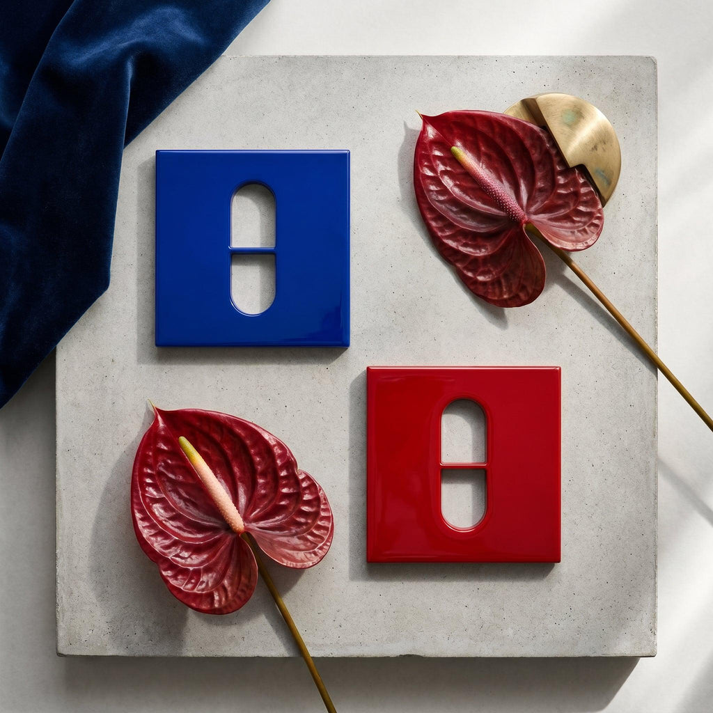 Two square light switch plates, one in glossy blue and one in glossy red, each featuring a central vertical pill-shaped cutout with a horizontal bar. Two dark red anthurium flowers with yellow spadices are placed alongside the plates.