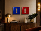 Two square wall art panels, one in deep blue and one in vibrant red, each featuring a centered, vertical pill-shaped cutout. The glossy surfaces reflect light, creating a clean, geometric aesthetic on the wall above a wooden console.