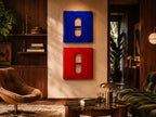 Two square wall art pieces mounted vertically, one in glossy blue and one in glossy red. Each piece features a central vertical cutout with a horizontal bar, creating a pill-like shape. The surfaces are smooth and reflective.