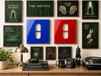 A gallery wall featuring framed posters with Matrix-themed imagery and quotes, alongside a glossy blue and a glossy red square wall plate, each with a central pill-shaped cutout, arranged above a desk with books, a lamp, and a figurine.