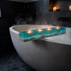 A rectangular bath tray made of clear resin with a turquoise blue tint, filled with small white pebbles at the base. The tray features four circular indentations, each holding a small lit tea light candle with a warm yellow flame.