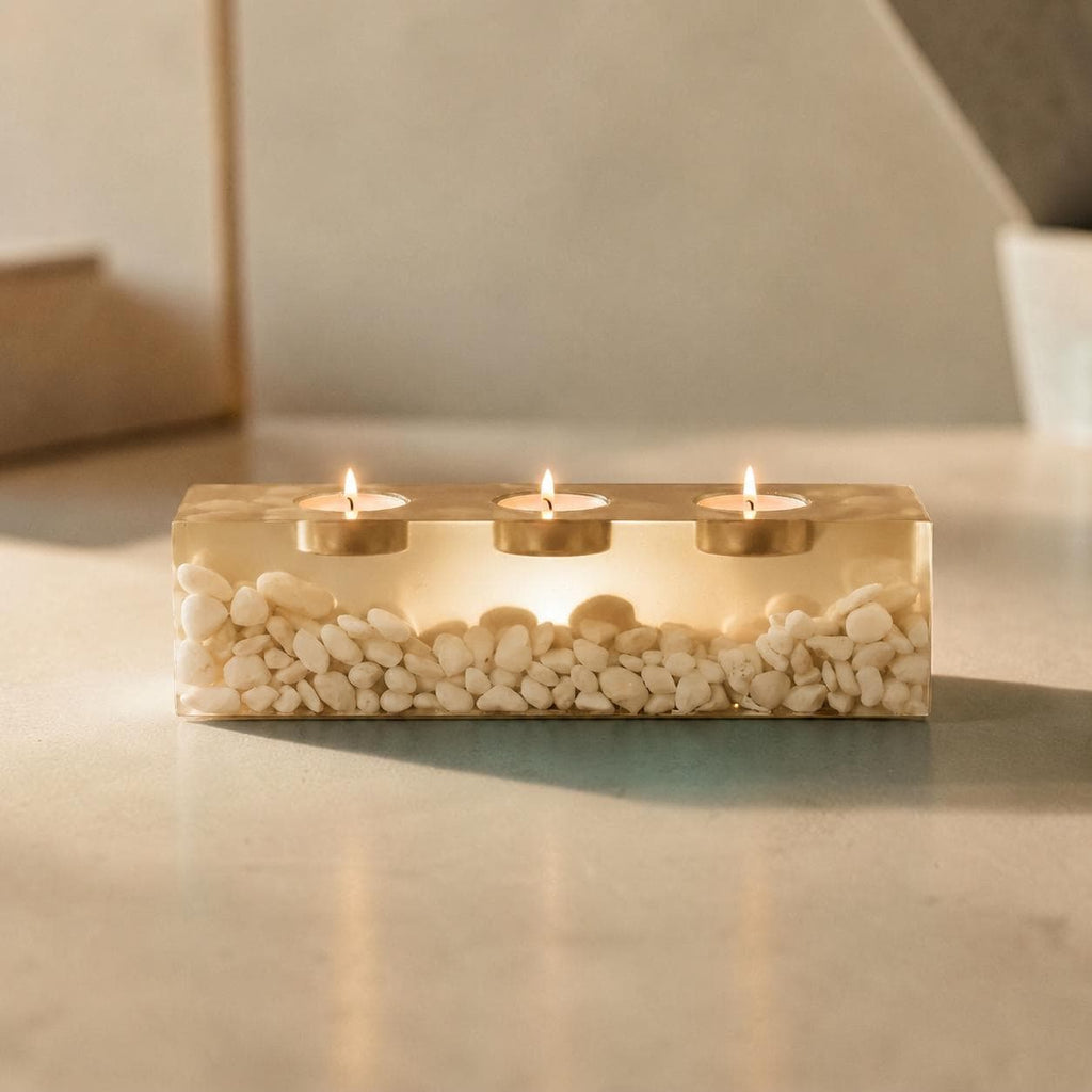 A rectangular clear resin candle holder containing numerous small white pebbles. The piece features three circular metal inserts, each holding a lit tea light candle with a visible flame, creating a warm glow through the resin.