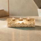 A rectangular clear resin candle holder containing numerous small white pebbles. The piece features three circular metal inserts, each holding a lit tea light candle with a visible flame, creating a warm glow through the resin.