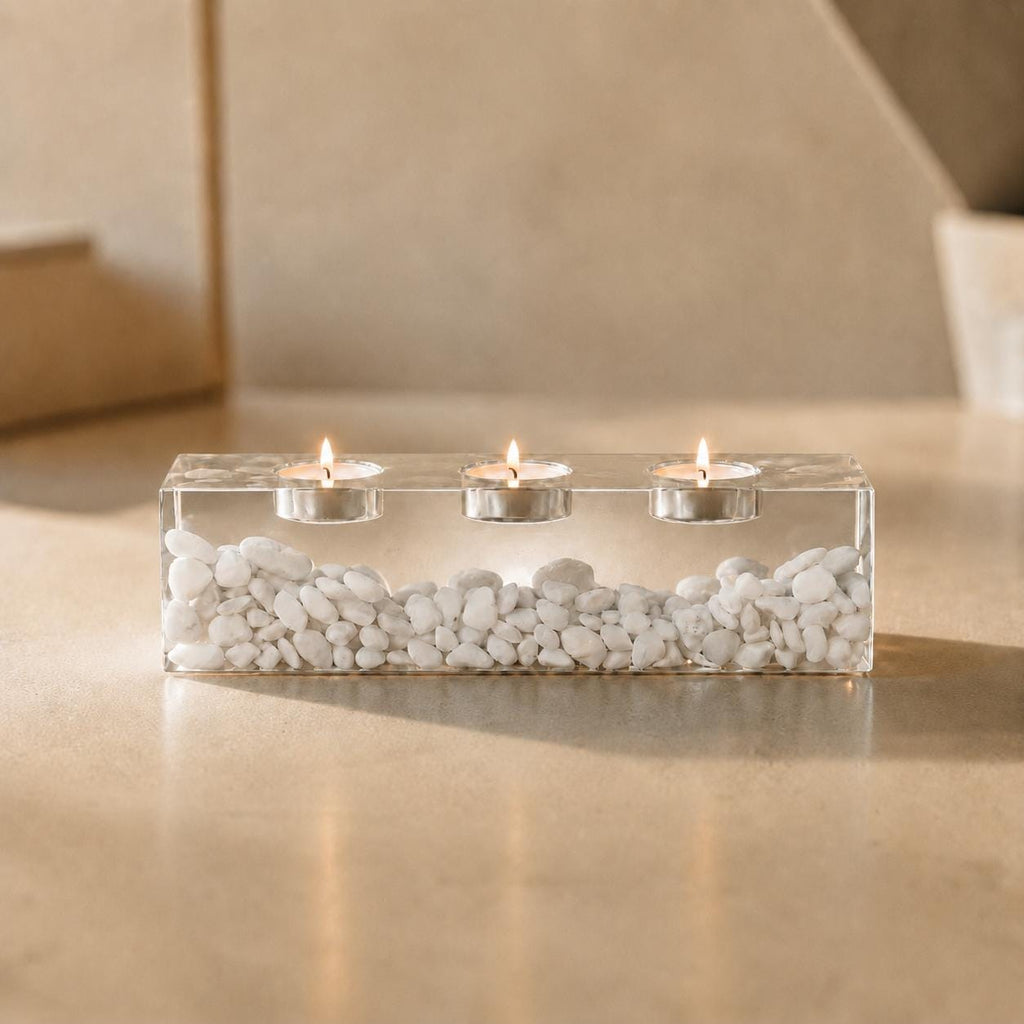 A rectangular clear resin candle holder containing a layer of small white stones at the base. The holder features three circular indentations along the top, each holding a lit tea light candle with a small metal casing and flame.