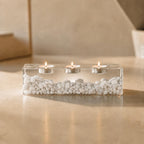 A rectangular clear resin candle holder containing a layer of small white stones at the base. The holder features three circular indentations along the top, each holding a lit tea light candle with a small metal casing and flame.