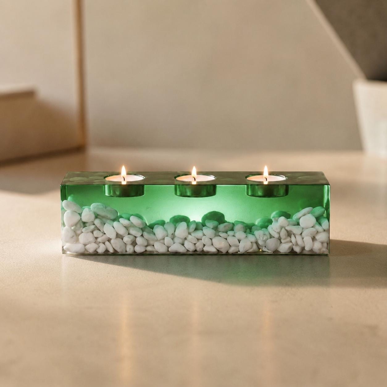 A rectangular green resin candle holder featuring three circular cutouts for tea light candles. The base of the holder is filled with small, smooth white stones embedded within the translucent green material, with three lit candles.