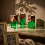 Three square candle holders made of natural brown wood and translucent green resin. Each holder features a dark wood top with a visible grain pattern and a smooth, vibrant green resin base. The tops have a circular cutout for candles.