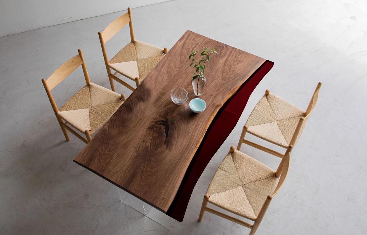 A rectangular dining table with a natural wood grain top and a dark red side edge, supported by clear acrylic legs. Four light-colored wooden chairs with woven seats are arranged around the table, which holds a glass vase and two bowls.