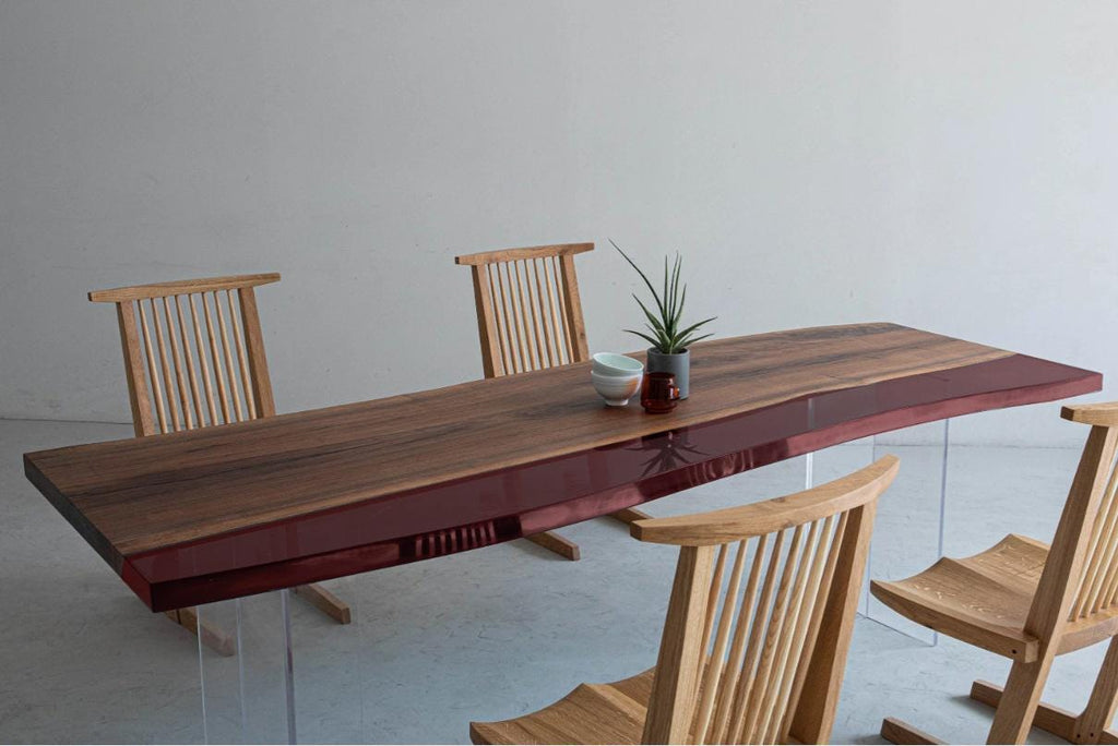 A rectangular dining table with a natural wood grain top and a deep red glossy edge, supported by clear acrylic legs. Three light-colored wooden chairs with vertical spindle backs are arranged around the table, which holds a small plant.