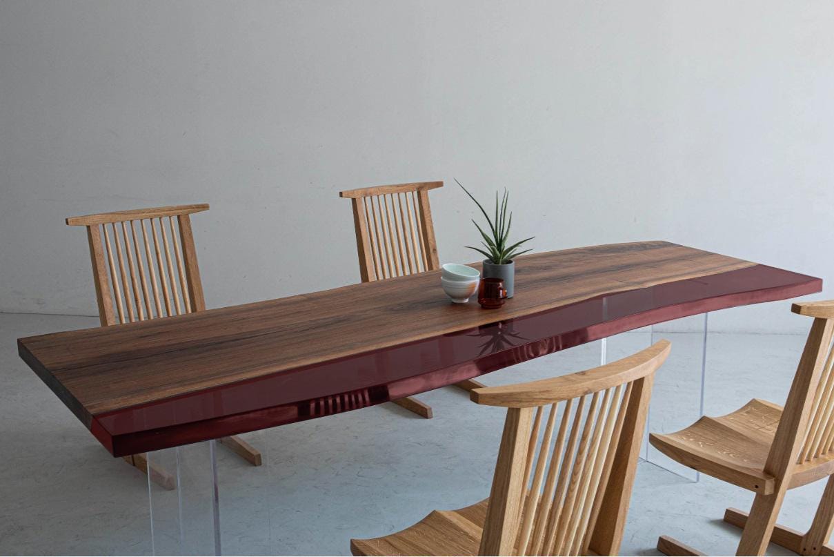 A rectangular dining table with a natural wood grain top and a deep red glossy edge, supported by clear acrylic legs. Three light-colored wooden chairs with vertical spindle backs are arranged around the table, which holds a small plant.