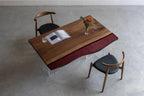 A rectangular dining table featuring a natural wood grain surface with a dark red glass accent panel along one side, supported by clear acrylic legs, accompanied by two wooden chairs with black upholstered seats and curved backrests.