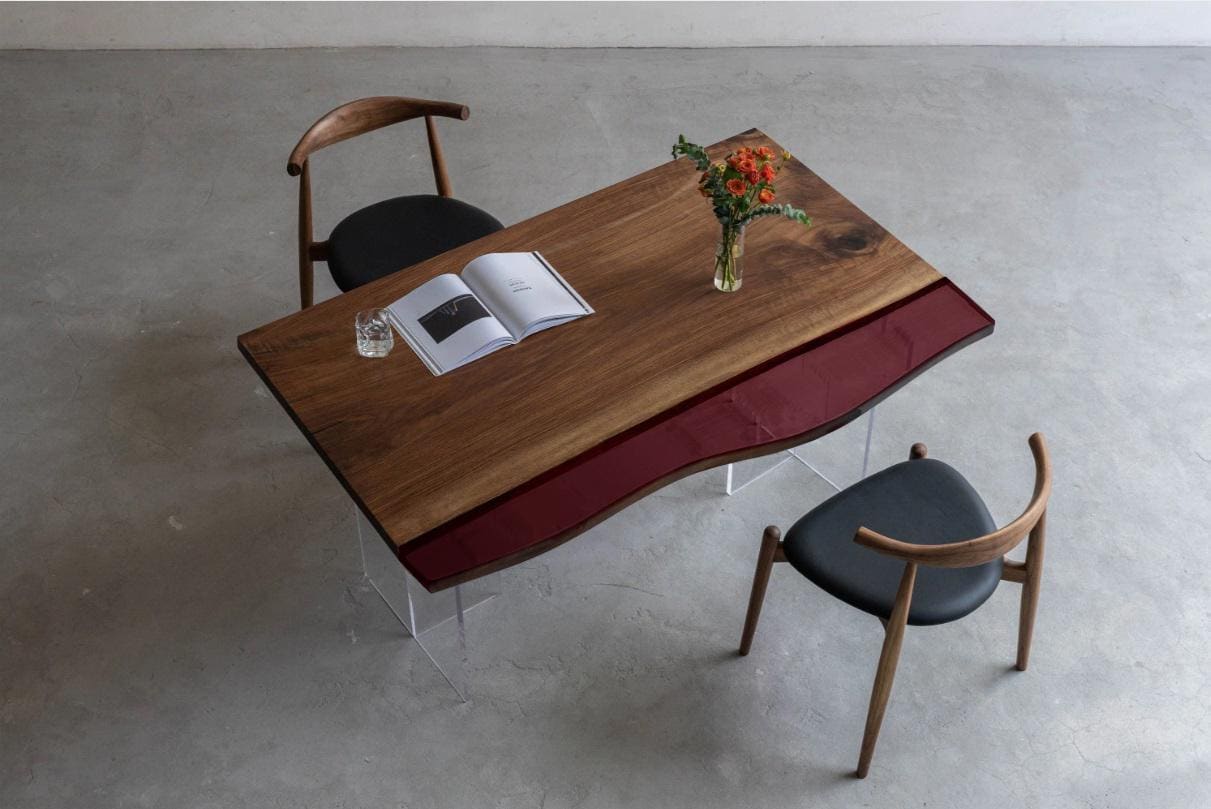 A rectangular dining table featuring a natural wood grain surface with a dark red glass accent panel along one side, supported by clear acrylic legs, accompanied by two wooden chairs with black upholstered seats and curved backrests.