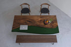A rectangular dining table featuring a natural live-edge wood slab paired with a solid dark green resin section. The set includes two wooden chairs with black circular seats and a matching wooden bench positioned below the table.