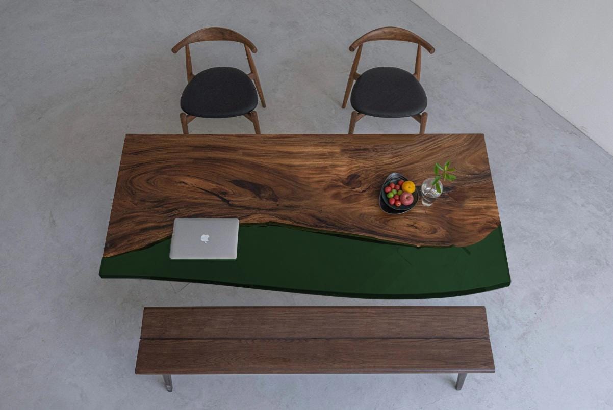 A rectangular dining table featuring a natural live-edge wood slab paired with a solid dark green resin section. The set includes two wooden chairs with black circular seats and a matching wooden bench positioned below the table.