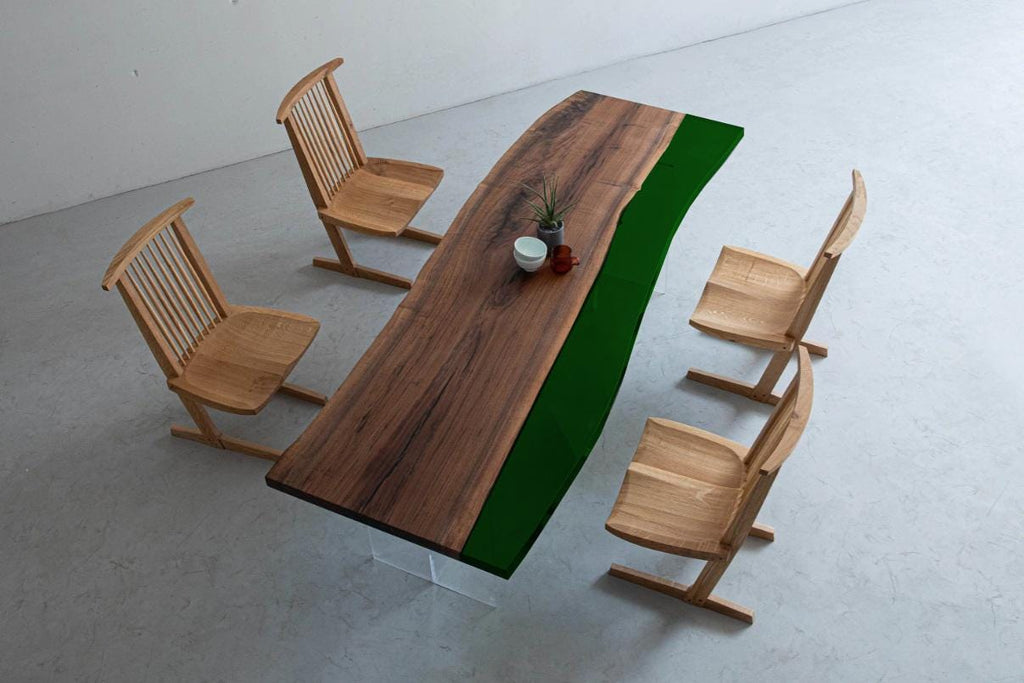 A dining set featuring a long rectangular table with a natural wood grain surface and a solid dark green resin edge, supported by clear acrylic legs, accompanied by four light-colored wooden chairs with slatted backrests.