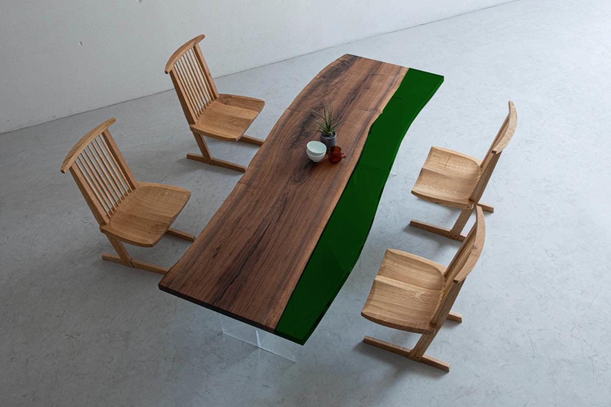 A dining set featuring a long rectangular table with a natural wood grain surface and a solid dark green resin edge, supported by clear acrylic legs, accompanied by four light-colored wooden chairs with slatted backrests.