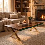 A rectangular coffee table with a natural wood grain top, a dark green resin edge, and gold-colored metal X-shaped legs. On the table sits a white ceramic mug, a stack of two books, a glass jar candle, and a small potted green plant.