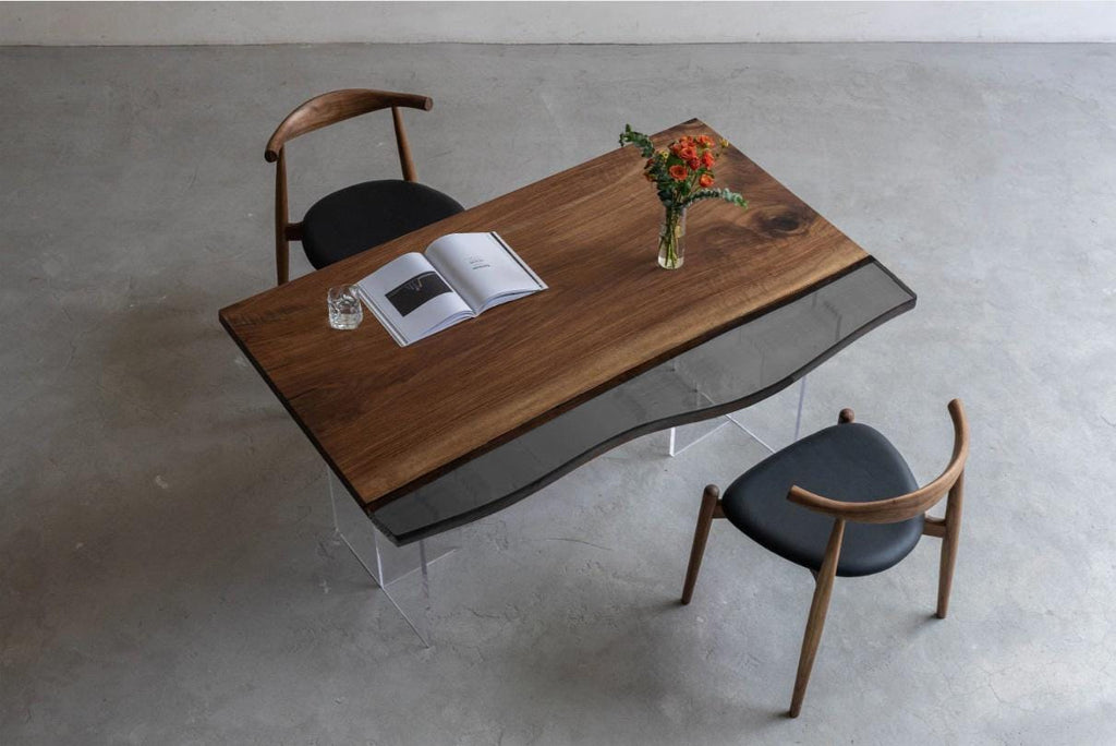 A rectangular dining table with a dark brown wood grain surface and a wavy, transparent glass edge section. The table is supported by clear acrylic legs and is accompanied by two wooden chairs with black upholstered seats.