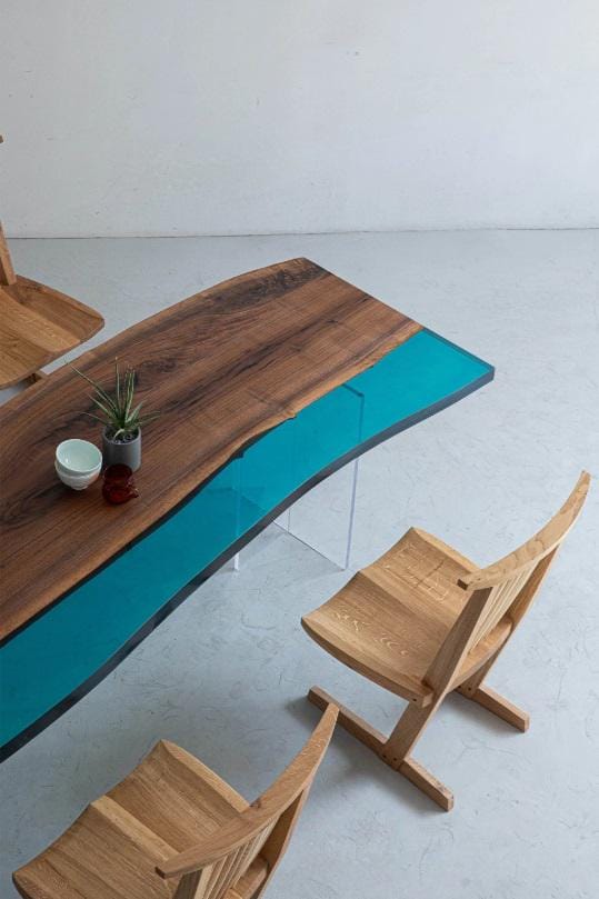 A dining table featuring a live-edge dark brown wood slab paired with a vibrant turquoise resin section, supported by clear acrylic legs. Two light-colored wooden chairs with curved seats and slatted backrests are positioned nearby.