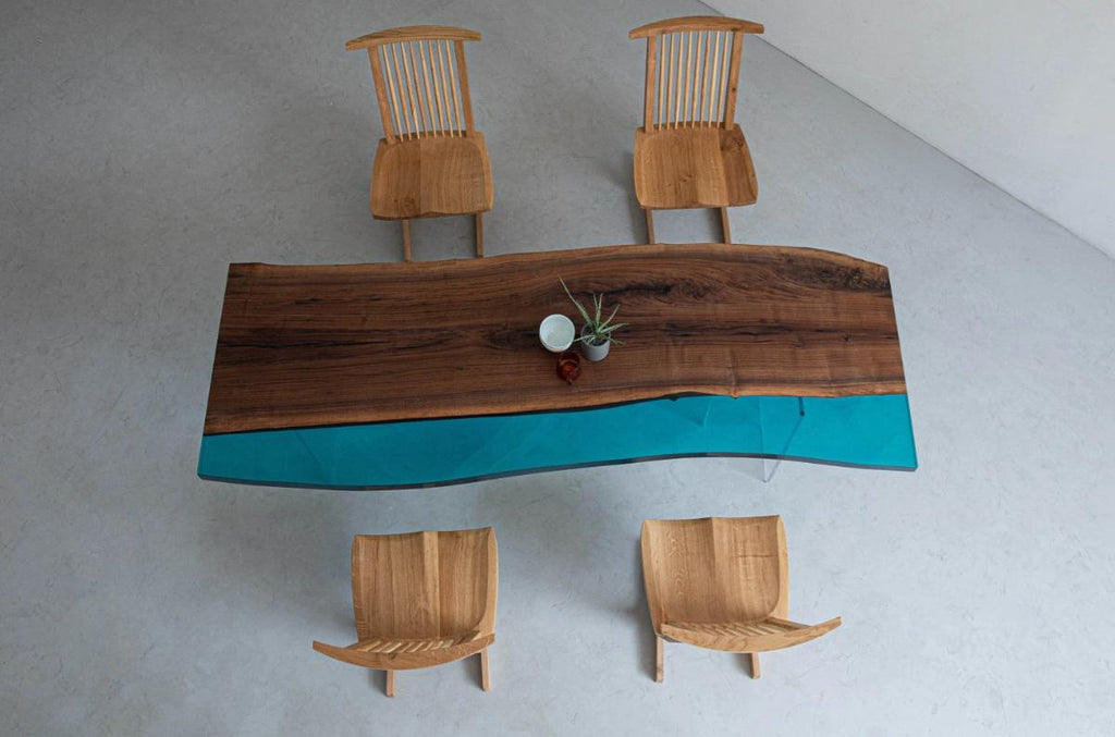 A rectangular dining table featuring a natural wood slab with a live edge and a translucent turquoise resin section, accompanied by four wooden spindle-back chairs. A small white bowl and a green plant sit on the table surface.