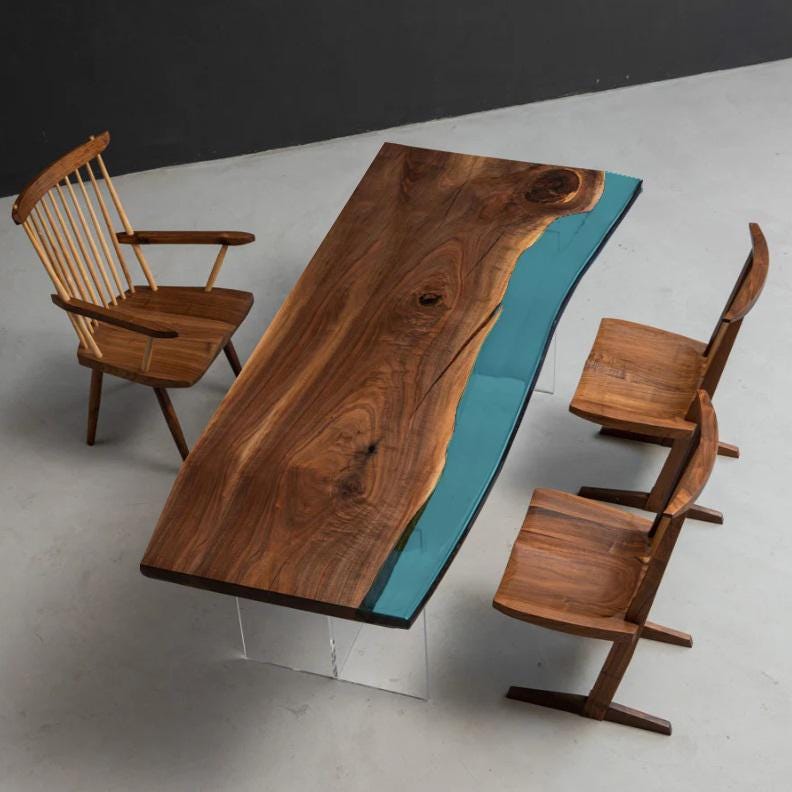 A rectangular dining table featuring a live-edge dark brown walnut wood slab with a central teal resin inlay, supported by clear acrylic legs, accompanied by three wooden chairs with varying back designs and angled legs.