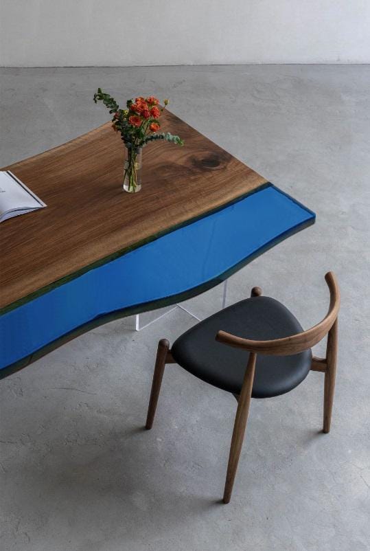 A dining table featuring a natural wood slab with a live edge and a smooth, solid blue resin inlay, paired with a wooden chair that has a curved backrest and a black upholstered seat. A small glass vase with flowers sits on top.