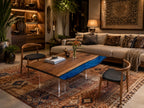 A rectangular coffee table featuring a natural wood grain surface with a wavy, translucent blue resin insert, supported by clear acrylic legs, accompanied by two wooden chairs with black upholstered seats on a patterned rug.