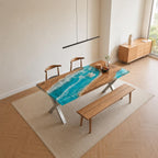 A rectangular dining table featuring a natural wood slab with a central river design made of blue and white resin that mimics ocean waves. The table has silver-colored metal X-shaped legs and is accompanied by two wooden chairs and a bench.