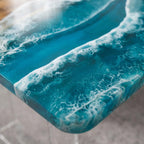 A rectangular table surface featuring a glossy resin finish with a deep teal and blue gradient. The surface displays white, swirling patterns that mimic the appearance of ocean waves crashing, with visible bubbles and depth.