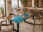 A rectangular dining table featuring a natural wood slab with a central river design made of vibrant blue and white resin that mimics ocean waves. The table is supported by black metal legs and is surrounded by wooden chairs with beige cushions.