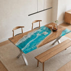 A rectangular dining table featuring a natural wood slab with a central river design made of bright turquoise resin that mimics ocean waves with white foam accents. The table has silver metal X-shaped legs and is paired with wooden chairs.