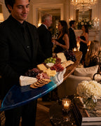 A long, surfboard-shaped wooden serving board featuring a deep blue and white resin wave design at one end. The board is arranged with an assortment of cheeses, cured meats, clusters of red and green grapes, and round crackers.