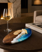 An oval serving board made of dark brown wood with a blue and white resin inlay resembling ocean waves. The board holds several round crackers and two pieces of cheese. A clear glass of white wine stands next to the board.