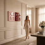 Two square wall art pieces featuring the word KISS in raised lettering. One piece is light pink and the other is deep red. The art is displayed on a paneled wall in a room with a beige slip dress, a marble table, and white flowers.