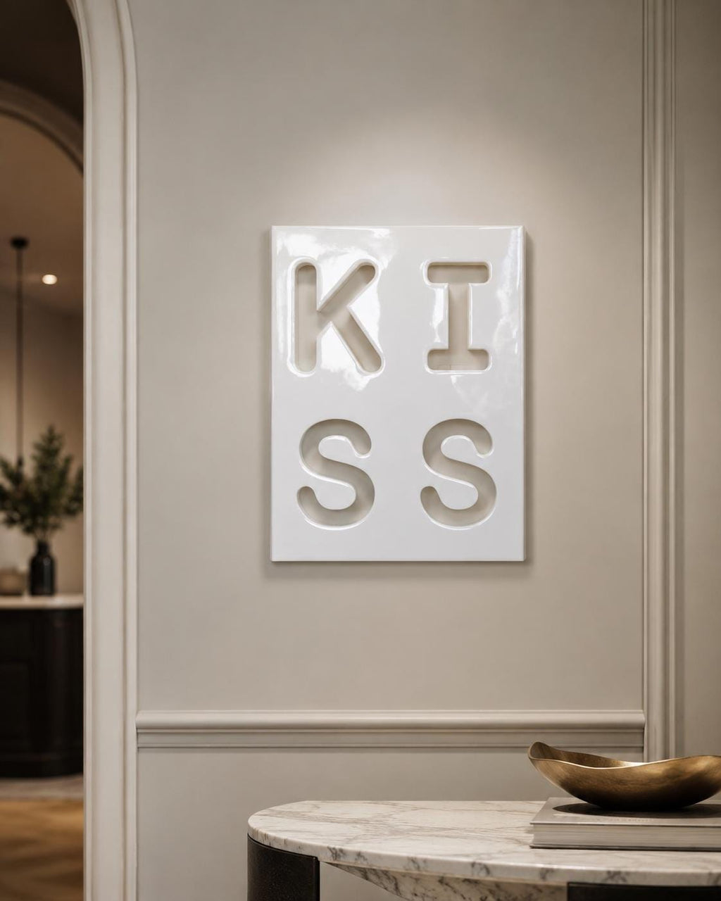 A rectangular white wall plaque featuring the word KISS embossed in a bold, recessed sans-serif font. The surface has a glossy, reflective finish, and the letters are arranged in a two-by-two grid format on the flat, smooth panel.