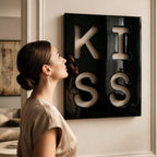 A square wall art piece featuring a glossy black surface with the word KISS cut out in large, bold, white-filled letters. The letters are arranged in a two-by-two grid, with K and I on top and S and S on the bottom.