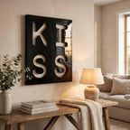 A square black wall art piece featuring the word KISS in large, bold, white cut-out letters. The glossy black surface surrounds the white lettering, creating a high-contrast design that is mounted on a light-colored wall.