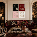 Four square wall art panels arranged in a grid, each featuring the word KISS in cut-out lettering. The panels are colored black, red, light pink, and white, displayed above a deep red velvet sofa with patterned throw pillows.