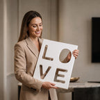 A rectangular white wall plaque featuring the word LOVE cut out in a bold, sans-serif font. The letters L, O, V, and E are arranged in a two-by-two grid, creating negative space through the solid white panel surface.