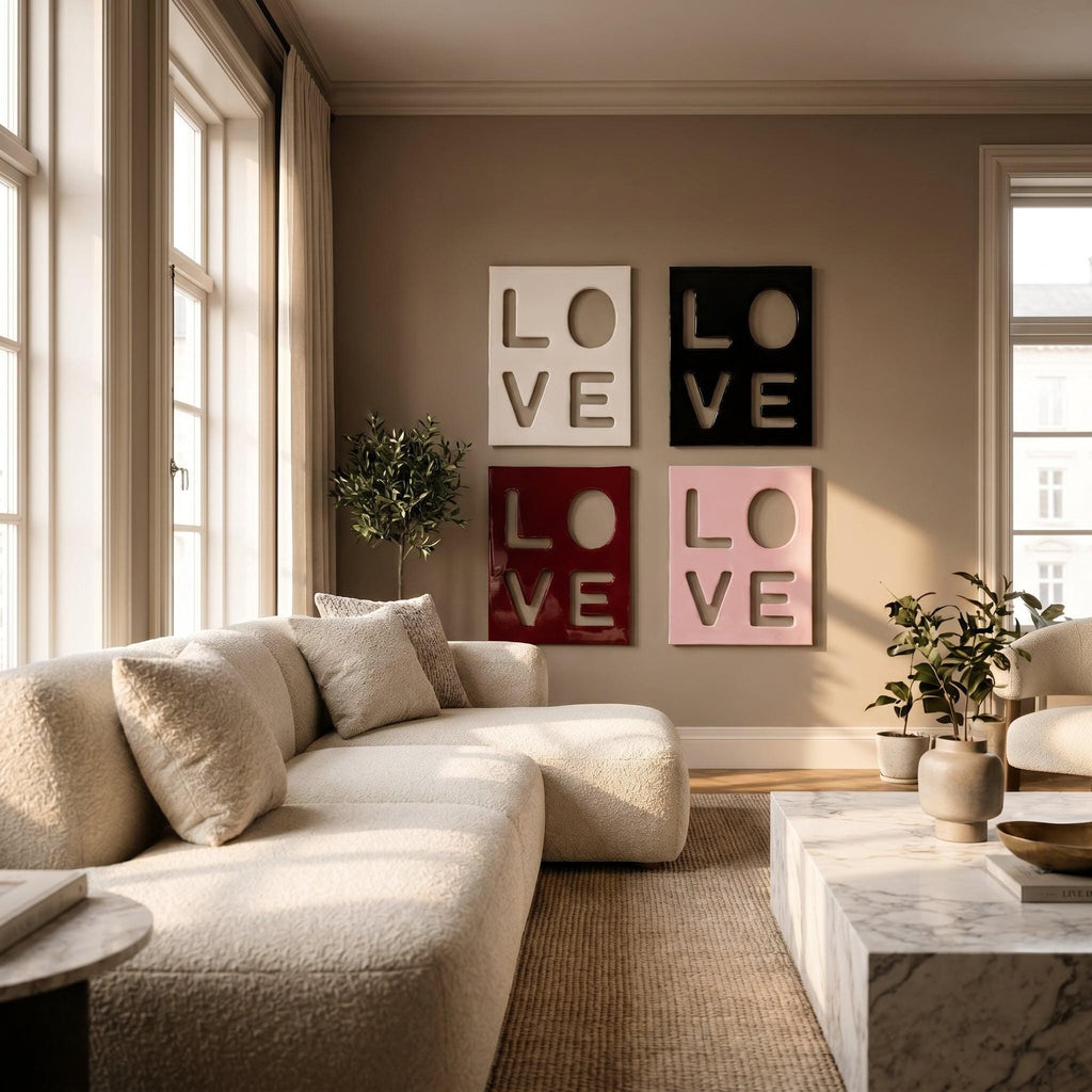 Four square wall art panels arranged in a grid, each featuring the word LOVE cut out in a bold, block-letter style. The panels are colored white, black, deep red, and light pink, displayed on a neutral-toned wall above a sofa.
