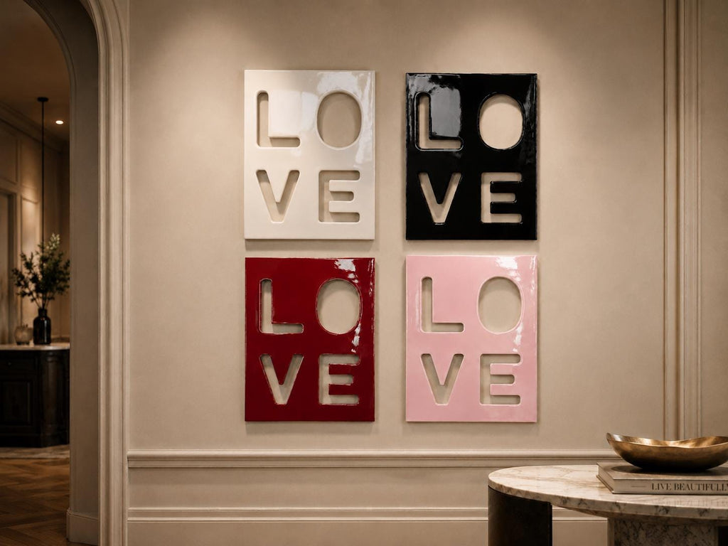 Four square wall art panels arranged in a grid, each featuring the word LOVE cut out in a bold, block-letter style. The panels are finished in a glossy glaze, with colors including white, black, deep red, and soft pastel pink.